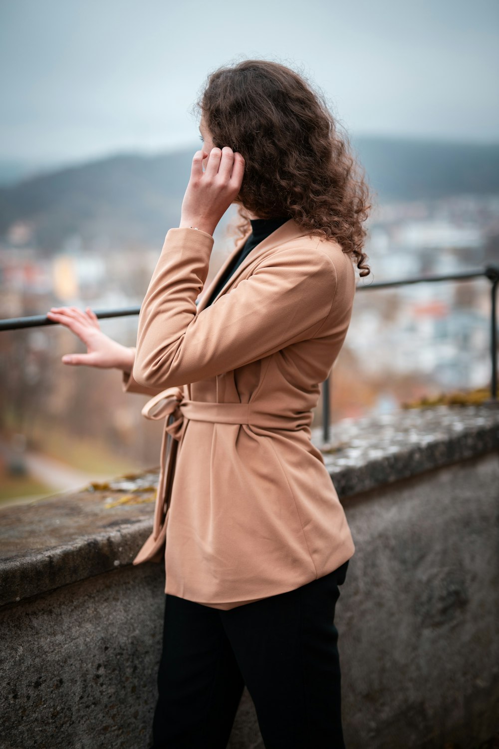 woman in brown coat standing on gray concrete wall during daytime