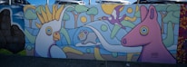 Brightly colored mural featuring stylized animals including a large bird, a smaller bird in flight, a sheep, and a horse. The background includes abstract trees and a sun. The artwork is playful with bold outlines and a cartoon-like appearance.