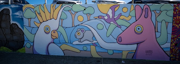 Brightly colored mural featuring stylized animals including a large bird, a smaller bird in flight, a sheep, and a horse. The background includes abstract trees and a sun. The artwork is playful with bold outlines and a cartoon-like appearance.