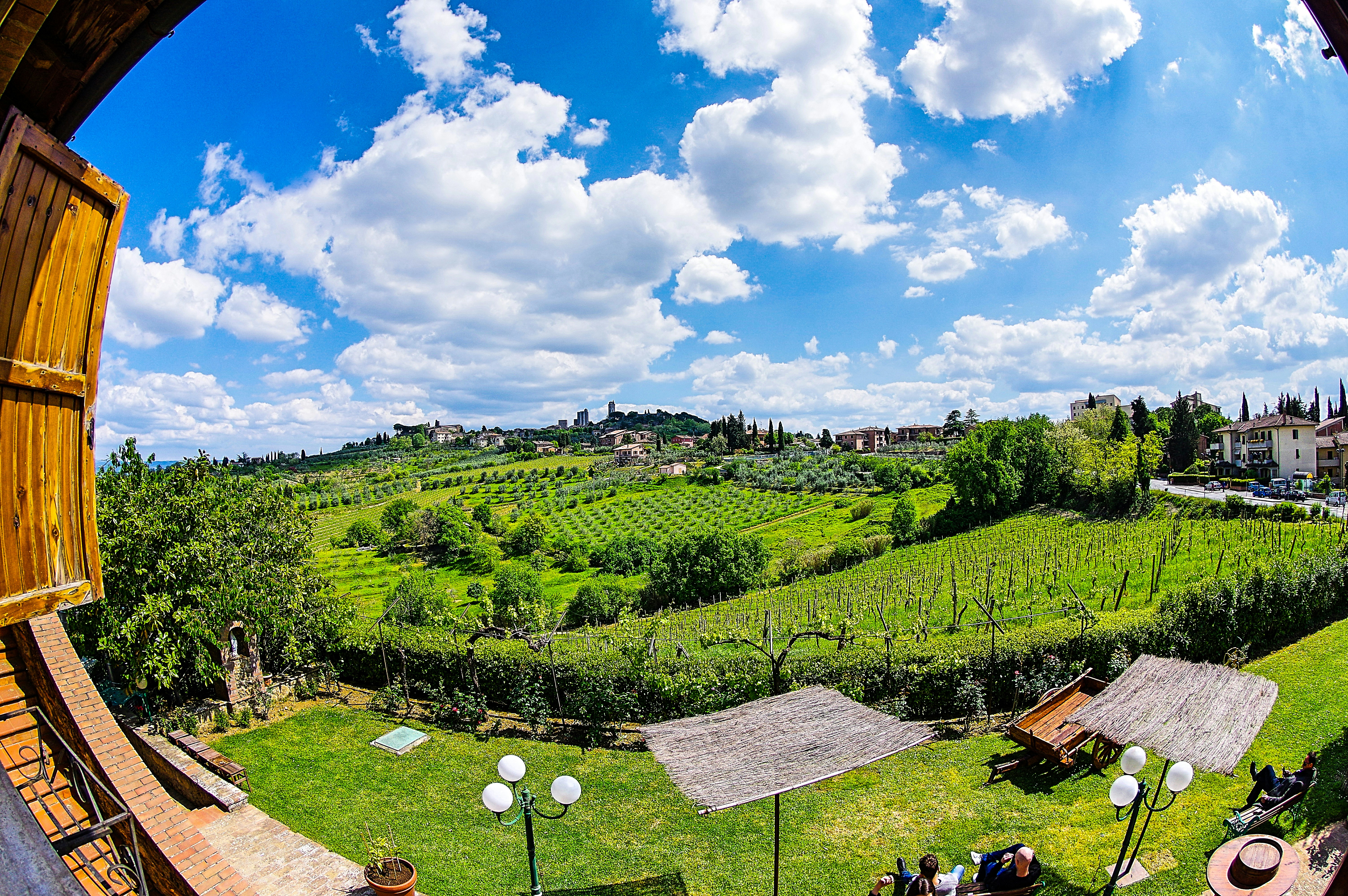 A picturesque countryside landscape features rolling green hills and lush vineyards under a bright blue sky with scattered white clouds. In the foreground, wooden structures, possibly pergolas, provide shade over tables and chairs on a well-manicured lawn. Traditional buildings can be seen on the hillside, creating a serene and pastoral setting.