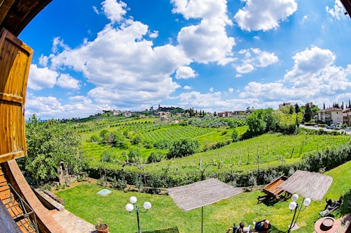 A picturesque countryside landscape features rolling green hills and lush vineyards under a bright blue sky with scattered white clouds. In the foreground, wooden structures, possibly pergolas, provide shade over tables and chairs on a well-manicured lawn. Traditional buildings can be seen on the hillside, creating a serene and pastoral setting.