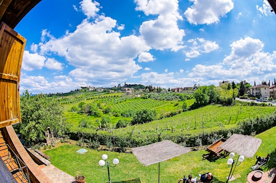 A picturesque countryside landscape features rolling green hills and lush vineyards under a bright blue sky with scattered white clouds. In the foreground, wooden structures, possibly pergolas, provide shade over tables and chairs on a well-manicured lawn. Traditional buildings can be seen on the hillside, creating a serene and pastoral setting.