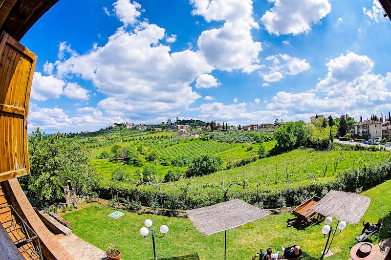 A picturesque countryside landscape features rolling green hills and lush vineyards under a bright blue sky with scattered white clouds. In the foreground, wooden structures, possibly pergolas, provide shade over tables and chairs on a well-manicured lawn. Traditional buildings can be seen on the hillside, creating a serene and pastoral setting.