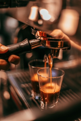 A close-up view of espresso being brewed into two small glass cups from a coffee machine. A hand is holding the portafilter, and the rich, dark liquid is flowing smoothly. The warm lighting enhances the inviting, aromatic ambiance typical of a cozy cafe setting.