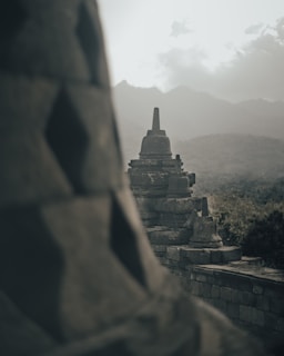 Ancient stone stupas stand prominently at the Borobudur Temple complex, with misty mountains in the background. The scene is serene and atmospheric, capturing the essence of a historical site shrouded in mystery.