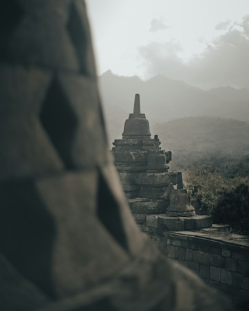 Ancient stone stupas stand prominently at the Borobudur Temple complex, with misty mountains in the background. The scene is serene and atmospheric, capturing the essence of a historical site shrouded in mystery.
