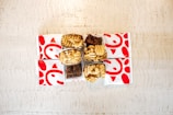 On a light wooden table, there are several fast-food items. The setup includes waffle fries in open boxes and square-shaped brownies. The arrangement is surrounded by paper bags with a distinctive red logo featuring stylized shapes.