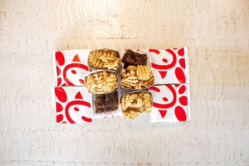 On a light wooden table, there are several fast-food items. The setup includes waffle fries in open boxes and square-shaped brownies. The arrangement is surrounded by paper bags with a distinctive red logo featuring stylized shapes.