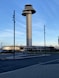 A tall, cylindrical control tower stands prominently against a clear blue sky with horizontal cloud streaks. Surrounding the tower is a modern building complex with large windows. The foreground features a paved road and sections of a sidewalk, with several tall lamp posts lining the street.