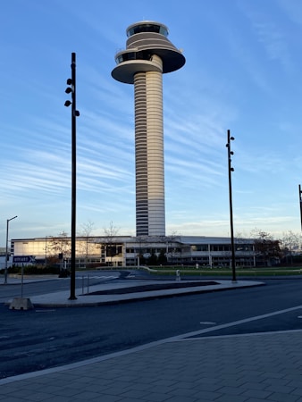 A tall, cylindrical control tower stands prominently against a clear blue sky with horizontal cloud streaks. Surrounding the tower is a modern building complex with large windows. The foreground features a paved road and sections of a sidewalk, with several tall lamp posts lining the street.