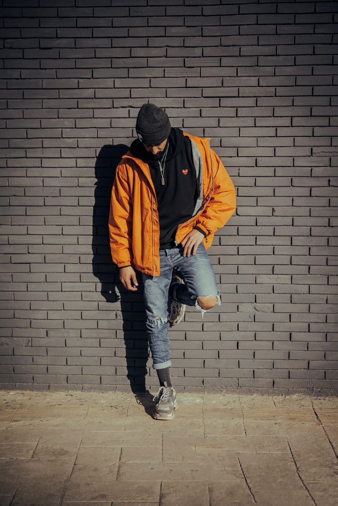 man in orange jacket and blue denim jeans standing beside brick wall