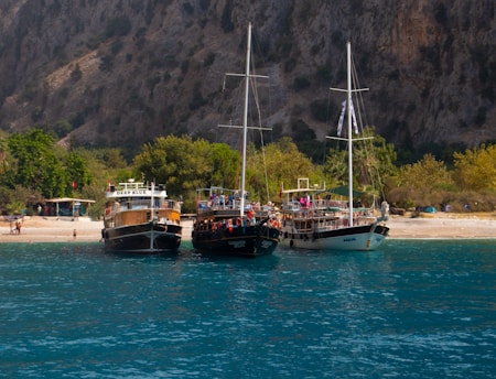 Three boats are anchored near a sandy beach with lush greenery and a rocky hillside in the background. The boats, named 'Deep Blue', 'Aqua', and another vessel, are moored side by side in the clear, calm water. People can be seen on and around the boats, likely enjoying leisure activities. The scene has a tranquil and inviting atmosphere, suggesting a popular spot for tourists or locals.