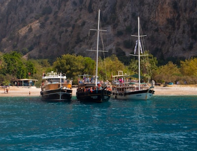 Three boats are anchored near a sandy beach with lush greenery and a rocky hillside in the background. The boats, named 'Deep Blue', 'Aqua', and another vessel, are moored side by side in the clear, calm water. People can be seen on and around the boats, likely enjoying leisure activities. The scene has a tranquil and inviting atmosphere, suggesting a popular spot for tourists or locals.