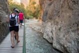 People walking along a narrow pathway beside a rocky gorge with clear stream water flowing below. Dense greenery is visible atop the canyon walls, with sunlight filtering through the trees.