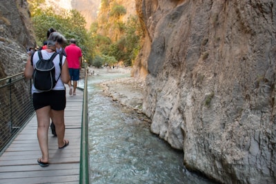 People walking along a narrow pathway beside a rocky gorge with clear stream water flowing below. Dense greenery is visible atop the canyon walls, with sunlight filtering through the trees.