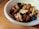 A white bowl filled with a mixture of nuts and dried fruits rests on a wooden surface. The nuts include almonds and walnuts, while the dried fruits appear to be dark in color, possibly raisins or similar. The combination creates a healthy and nutritious snack.