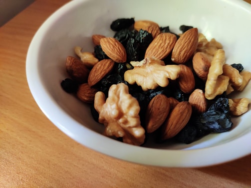 A white bowl filled with a mixture of nuts and dried fruits rests on a wooden surface. The nuts include almonds and walnuts, while the dried fruits appear to be dark in color, possibly raisins or similar. The combination creates a healthy and nutritious snack.