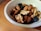 A white bowl filled with a mixture of nuts and dried fruits rests on a wooden surface. The nuts include almonds and walnuts, while the dried fruits appear to be dark in color, possibly raisins or similar. The combination creates a healthy and nutritious snack.