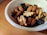 A white bowl filled with a mixture of nuts and dried fruits rests on a wooden surface. The nuts include almonds and walnuts, while the dried fruits appear to be dark in color, possibly raisins or similar. The combination creates a healthy and nutritious snack.