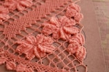 Detailed crochet work in pink, showcasing intricate floral patterns on a grid of loops. The flowers are prominently featured in a dense arrangement, highlighting craftsmanship and texture against a neutral background.
