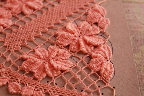 Detailed crochet work in pink, showcasing intricate floral patterns on a grid of loops. The flowers are prominently featured in a dense arrangement, highlighting craftsmanship and texture against a neutral background.