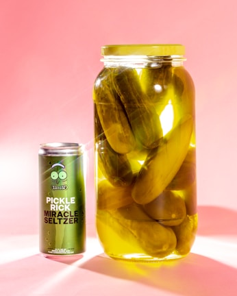 A large jar of pickles is placed next to a green can of seltzer water labeled 'Pickle Rick Miracle Seltzer.' The jar and the can are set against a soft pink background, and the pickles in the jar are submerged in yellowish pickle brine.