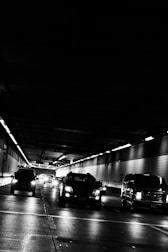 Vehicles are driving through an urban tunnel with overhead lights casting reflections on the road surface. The environment appears dimly lit, emphasizing the contrast between the vehicles' headlights and the dark surroundings.
