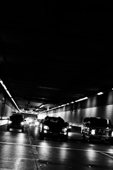 Vehicles are driving through an urban tunnel with overhead lights casting reflections on the road surface. The environment appears dimly lit, emphasizing the contrast between the vehicles' headlights and the dark surroundings.