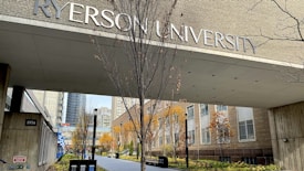 An entrance to a university building with large lettering displaying the university's name. The pathway is lined with trees that have autumn foliage, and surrounding buildings are visible in the background.