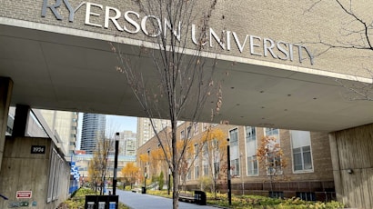 An entrance to a university building with large lettering displaying the university's name. The pathway is lined with trees that have autumn foliage, and surrounding buildings are visible in the background.