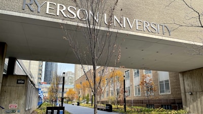 An entrance to a university building with large lettering displaying the university's name. The pathway is lined with trees that have autumn foliage, and surrounding buildings are visible in the background.