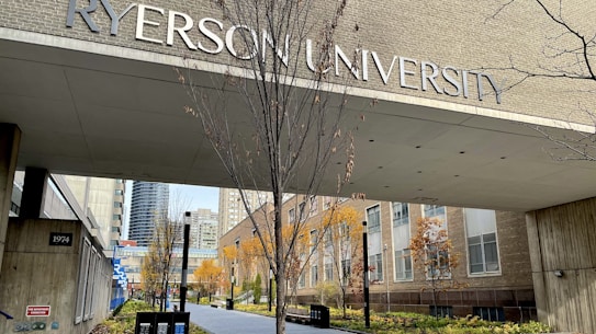 An entrance to a university building with large lettering displaying the university's name. The pathway is lined with trees that have autumn foliage, and surrounding buildings are visible in the background.