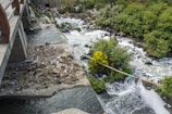 A small river or stream with foamy water flows under a concrete bridge. On the riverbank, there is a large accumulation of trash and debris. Dense foliage and green bushes grow along the river, providing a stark contrast to the polluted area.