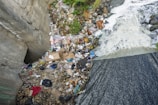 A polluted riverbank is littered with a significant amount of trash, including discarded boxes, plastic bags, and various debris. The water is flowing over the edge, creating white foam, with trash scattered around the bank, indicating environmental pollution and neglect.