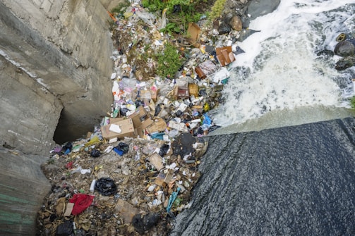 A polluted riverbank is littered with a significant amount of trash, including discarded boxes, plastic bags, and various debris. The water is flowing over the edge, creating white foam, with trash scattered around the bank, indicating environmental pollution and neglect.
