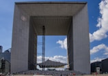 A large, modern architectural structure resembling a cube with an open center. The edifice features a grid-like pattern on its surfaces and is surrounded by a clear blue sky with scattered clouds. Numerous people are visible at the base, indicating the building's vast scale.