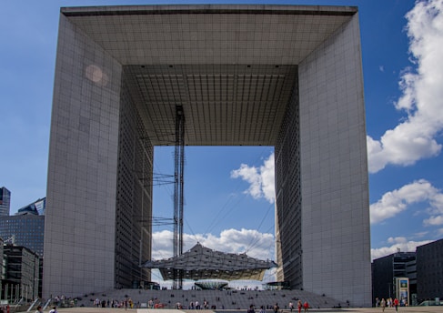 A large, modern architectural structure resembling a cube with an open center. The edifice features a grid-like pattern on its surfaces and is surrounded by a clear blue sky with scattered clouds. Numerous people are visible at the base, indicating the building's vast scale.