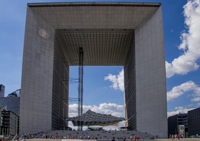 A large, modern architectural structure resembling a cube with an open center. The edifice features a grid-like pattern on its surfaces and is surrounded by a clear blue sky with scattered clouds. Numerous people are visible at the base, indicating the building's vast scale.