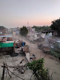 A rural or semi-urban area features small buildings with metal and brick structures. Dirt roads are visible, and there is a horse-drawn cart along with a motorcycle parked nearby. Trees and foliage are present, and the sky shows a gradient of colors, suggesting either sunrise or sunset. The area seems quiet and dusty, with some people walking in the distance.