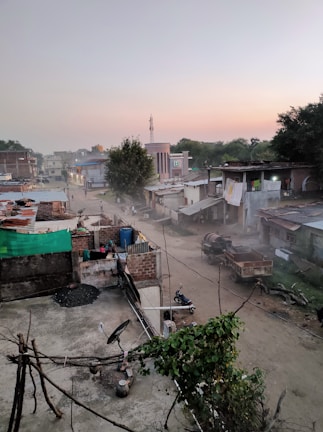 A rural or semi-urban area features small buildings with metal and brick structures. Dirt roads are visible, and there is a horse-drawn cart along with a motorcycle parked nearby. Trees and foliage are present, and the sky shows a gradient of colors, suggesting either sunrise or sunset. The area seems quiet and dusty, with some people walking in the distance.