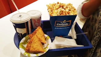 A tray containing two cups of soft drinks with straws, a large blue box of popcorn, and a plate with three samosas arranged neatly. The tray is blue and is placed on a white surface. A napkin is also visible next to the food items.