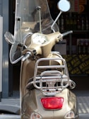 A beige scooter is parked with a clear windshield and two mirrors attached. The handlebars and the dashboard with the speedometer are visible. The scooter is located near another vehicle's open trunk where various bottles and boxes are stored.
