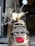 A beige scooter is parked with a clear windshield and two mirrors attached. The handlebars and the dashboard with the speedometer are visible. The scooter is located near another vehicle's open trunk where various bottles and boxes are stored.
