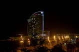 A tall, modern hotel building is illuminated at night, with its distinctive curved structure resembling a sail. The area around the hotel is lit with streetlights, creating a warm glow contrasted against the dark night sky. The hotel is located near a waterfront, with some trees visible in the foreground.
