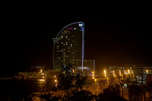A tall, modern hotel building is illuminated at night, with its distinctive curved structure resembling a sail. The area around the hotel is lit with streetlights, creating a warm glow contrasted against the dark night sky. The hotel is located near a waterfront, with some trees visible in the foreground.