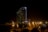 A tall, modern hotel building is illuminated at night, with its distinctive curved structure resembling a sail. The area around the hotel is lit with streetlights, creating a warm glow contrasted against the dark night sky. The hotel is located near a waterfront, with some trees visible in the foreground.