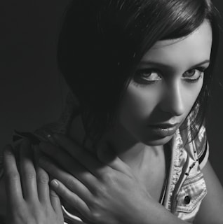 Black and white portrait of a woman with dark hair, looking intently at the camera. Her hands are crossed over her chest, adding a sense of introspection. The lighting is dramatic, casting shadows and highlighting her facial features.