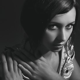 Black and white portrait of a woman with dark hair, looking intently at the camera. Her hands are crossed over her chest, adding a sense of introspection. The lighting is dramatic, casting shadows and highlighting her facial features.