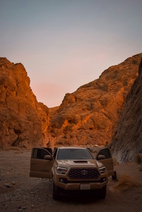 A rugged desert landscape at sunset with rocky hills glowing in a warm golden hue. In the foreground, a beige pickup truck with its doors open is parked on the sandy ground.