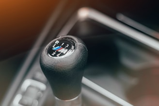 A close-up of a black manual gear shift knob with the M logo, surrounded by a metallic gear panel. The numbers and letters are clearly visible, indicating the gear positions.